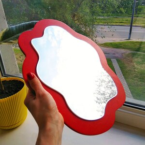Pu&ograve; includere: Uno specchio da mano rosso di forma irregolare con una superficie riflettente argentata. Lo specchio &egrave; tenuto da una mano.