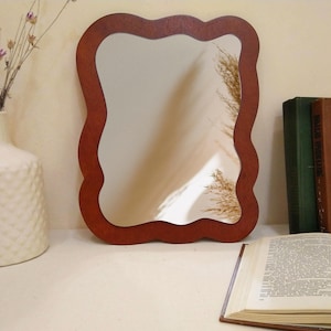 Puede incluir: Espejo decorativo con marco de madera marrón oscuro ondulado. El espejo refleja una pared de color claro y un toque de plantas secas. También se ven un jarrón blanco con flores secas, libros y un libro abierto.