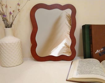 May include: A decorative mirror with a wavy, dark brown wooden frame. The mirror reflects a light-colored wall and a hint of dried plants. A white vase with dried flowers, books, and an open book are also visible.