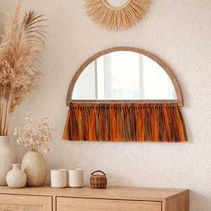 May include: A half-moon shaped mirror with a wooden frame and a fringe of orange and brown fabric. The mirror is mounted on a textured white wall. Decorative vases and a small basket sit on a wooden dresser below.