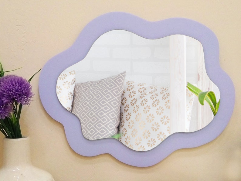 May include: A decorative wall mirror with an unusual, organic shape. The mirror has a soft, lavender-coloured frame. The reflection shows pillows with geometric and floral patterns. A vase with purple flowers is visible on the left.