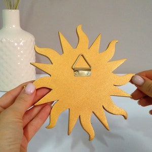 May include: Gold sun-shaped wall decor with a hanging hook on the back. The sun has a textured surface and is made of wood.