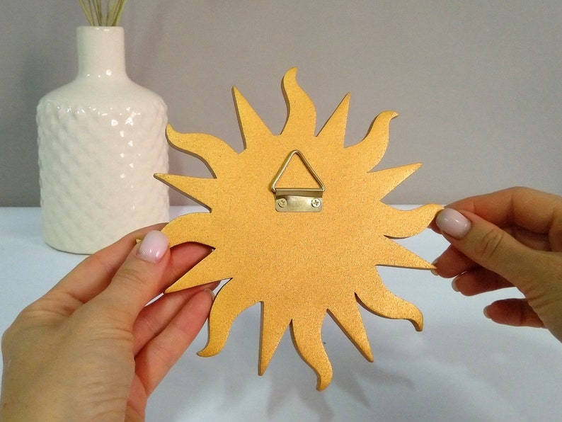 May include: Gold sun-shaped wall decor with a hanging hook on the back. The sun has a textured surface and is made of wood.