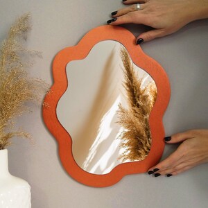 May include: A coral-colored, wavy-edged mirror hangs on a gray wall. The mirror reflects a dried flower arrangement and a glimpse of a room.