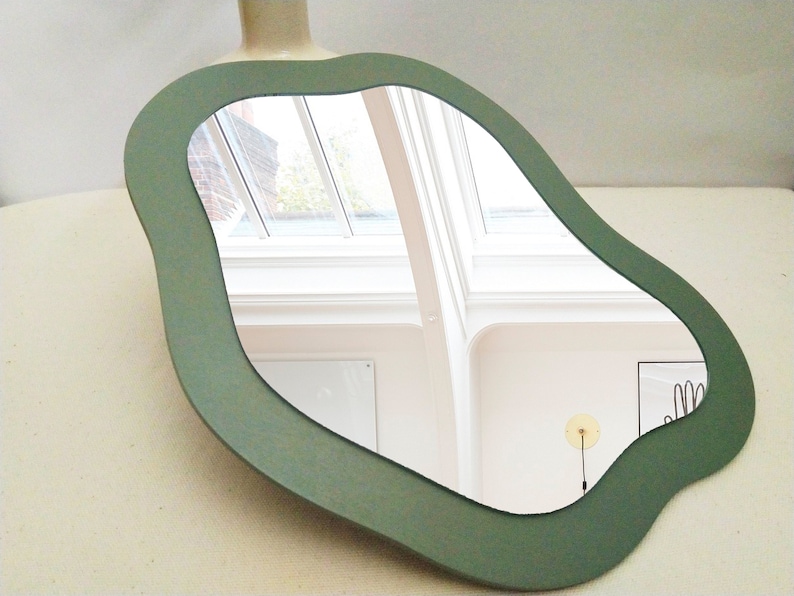 May include: A unique, sage green mirror with an organic, flowing shape. The mirror reflects a bright room with skylights. The frame is a solid, muted green, adding a touch of modern art to any space.
