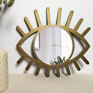 K&ouml;nnte beinhalten: Ein goldfarbener Wandspiegel in Augenform mit einer kreisf&ouml;rmigen Mitte und strahlenf&ouml;rmigen Holzlatten, die Wimpern nachahmen. Der Rahmen hat eine matte Oberfl&auml;che und umschlie&szlig;t eine klare, reflektierende Glasfl&auml;che mit Raumspiegelung.