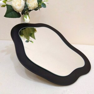 May include: A black, irregularly shaped mirror with a reflective surface. The mirror has a dark frame and is placed on a white surface. A vase of white flowers is in the background.