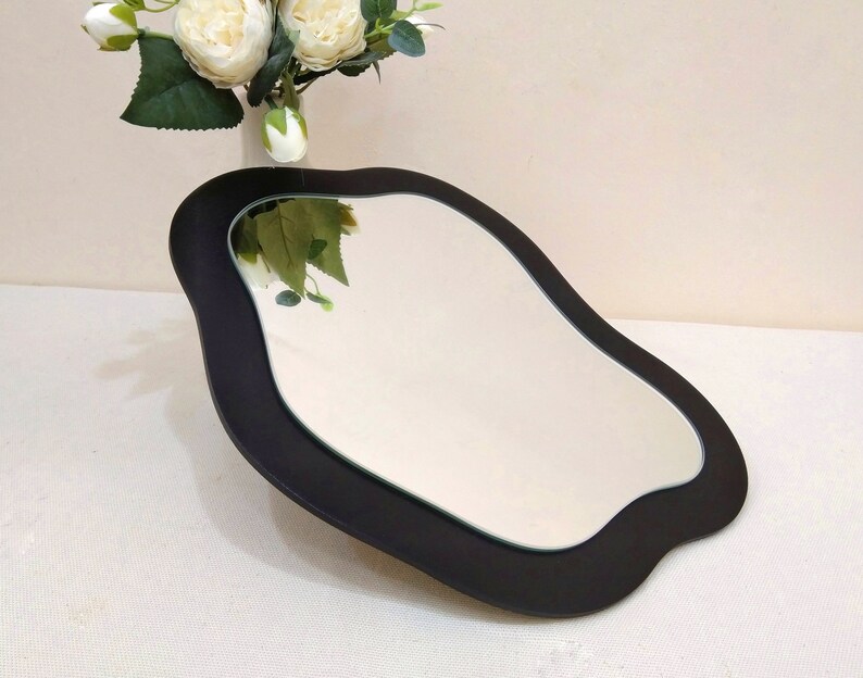 May include: A black, irregularly shaped mirror with a reflective surface. The mirror has a dark frame and is placed on a white surface. A vase of white flowers is in the background.