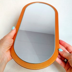 May include: A small, oval-shaped mirror with a gold-colored frame. The mirror is held in the hands of a person.