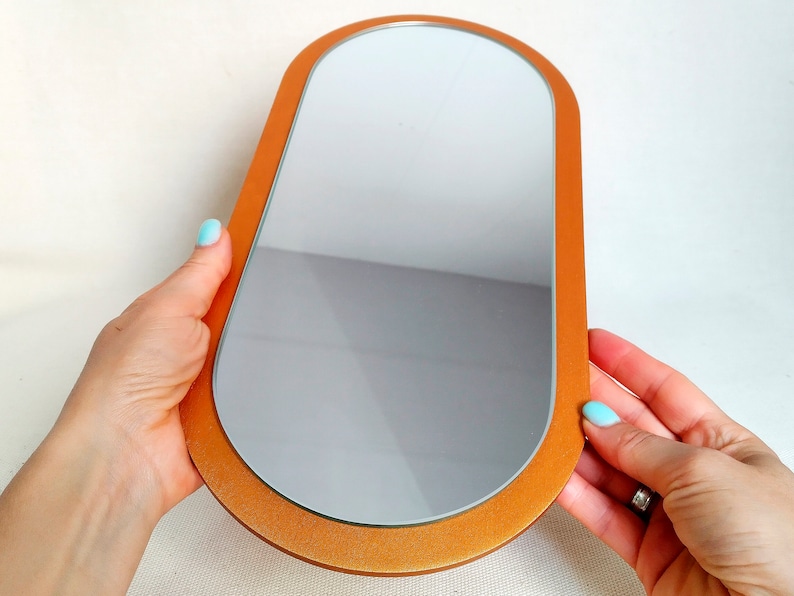 May include: A small, oval-shaped mirror with a gold-colored frame. The mirror is held in the hands of a person.
