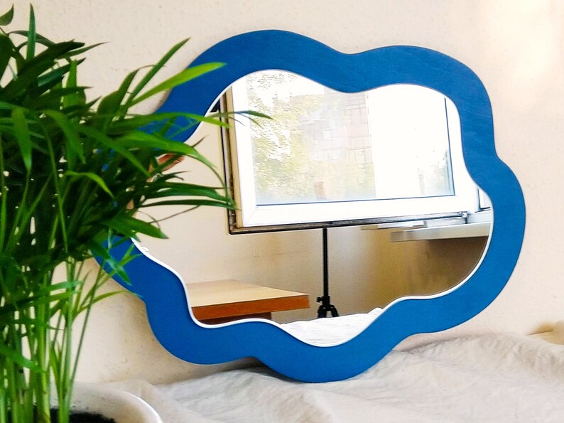 May include: A cloud-shaped mirror with a vibrant blue frame and a white inner border. The mirror reflects a window and a table. A green plant is in the foreground. The mirror is a decorative wall hanging.