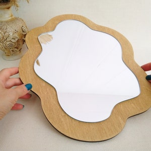 Puede incluir: Un espejo de madera con una forma &uacute;nica e irregular. El espejo tiene un marco de madera marr&oacute;n claro y una superficie de vidrio transparente.