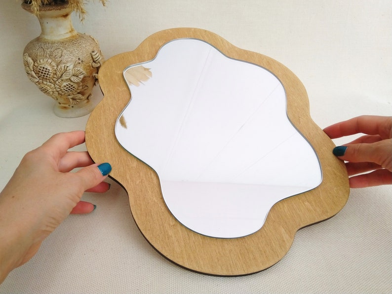 Puede incluir: Un espejo de madera con una forma &uacute;nica e irregular. El espejo tiene un marco de madera marr&oacute;n claro y una superficie de vidrio transparente.