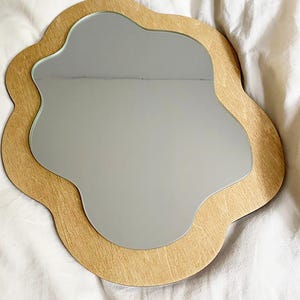 May include: A wooden framed mirror with a wavy, organic shape. The mirror is a light brown color and has a smooth, reflective surface.