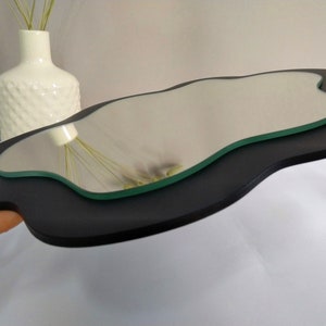 Puede incluir: Un marco de madera negro de forma irregular con un espejo de vidrio transparente ondulado en el interior. El espejo refleja un jarr&oacute;n blanco con flores secas.