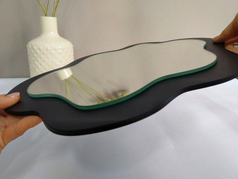 Puede incluir: Un marco de madera negro de forma irregular con un espejo de vidrio transparente ondulado en el interior. El espejo refleja un jarr&oacute;n blanco con flores secas.