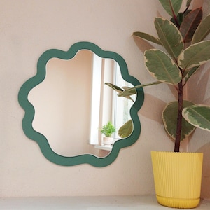 Puede incluir: Un espejo de pared decorativo con un marco único en forma de nube ondulada. El marco del espejo es de color verde apagado y refleja una habitación con una ventana y una pequeña planta. Una gran planta en maceta con hojas verdes y blancas está cerca.