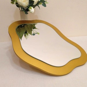 May include: A decorative mirror with a unique, flowing shape. The mirror has a gold-colored frame and a reflective surface. The design is modern and artistic, suitable for home decor. The mirror is placed on a white surface.