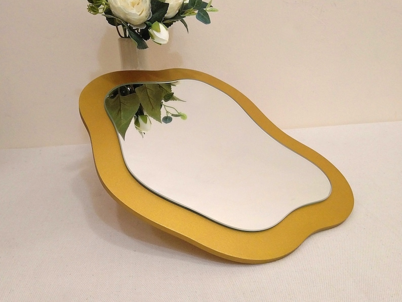 May include: A decorative mirror with a unique, flowing shape. The mirror has a gold-colored frame and a reflective surface. The design is modern and artistic, suitable for home decor. The mirror is placed on a white surface.