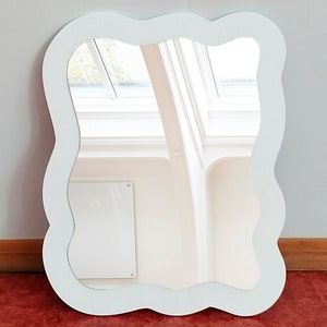 May include: A white, cloud-shaped mirror with a wavy edge. The mirror reflects a window and interior details. The frame is mounted on a white wall above a red floor. The mirror's dimensions are in centimetres.
