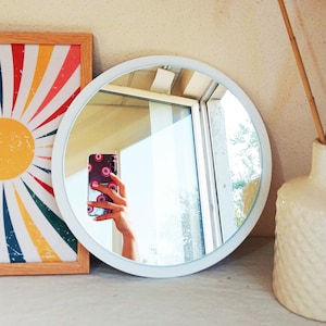 Op de afbeelding: Een ronde spiegel met een wit frame die een hand met een telefoon met een patroonhoesje weerspiegelt. Een kleurrijke zonsopgang kunstprint in een houten lijst staat links. Een witte, getextureerde vaas met gedroogde stelen staat rechts.