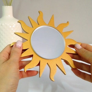 May include: A gold sun-shaped mirror with a round reflective surface. The mirror is held in a person's hands.