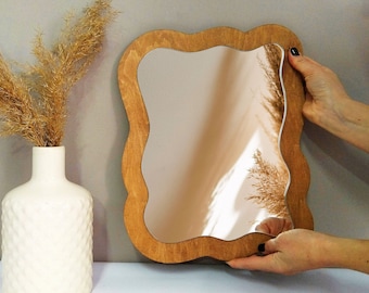 May include: A wavy wooden frame mirror with a light brown wood finish. The mirror is reflecting a bouquet of dried pampas grass.