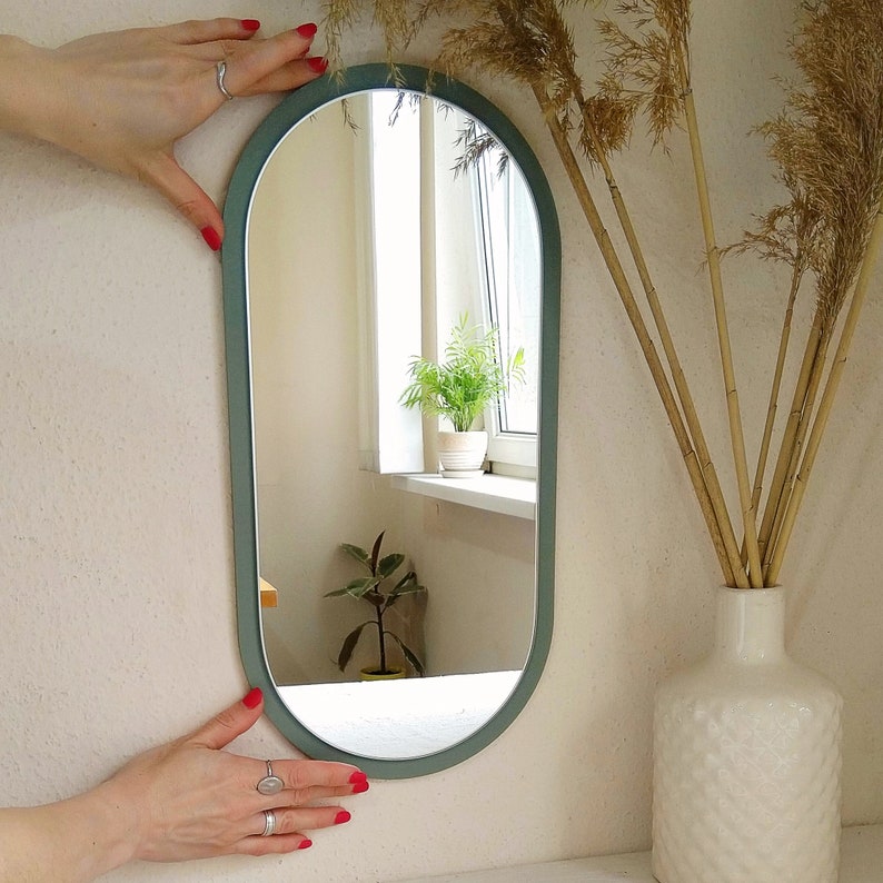 May include: A light green oval-shaped wall mirror with a rounded top and bottom. The mirror is reflecting a potted plant and a window.