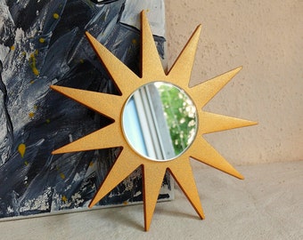 May include: A gold-colored starburst mirror with a circular mirror in the center. The starburst design has pointed rays extending outwards. The mirror reflects a view of a window and greenery. The mirror is placed in front of a painting.