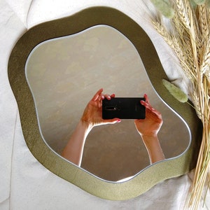 May include: An irregularly shaped mirror with a gold-colored frame. The mirror reflects a person holding a black smartphone. Dried wheat and green decorative plants are arranged to the right of the mirror, against a white background.