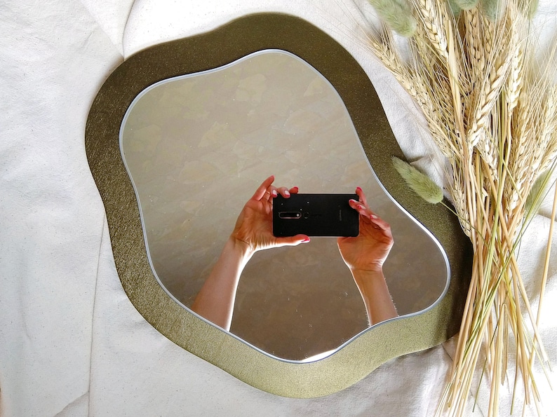May include: An irregularly shaped mirror with a gold-colored frame. The mirror reflects a person holding a black smartphone. Dried wheat and green decorative plants are arranged to the right of the mirror, against a white background.