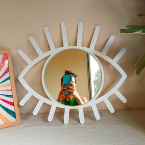 K&ouml;nnte beinhalten: Ein wei&szlig;er Wandspiegel in Augenform mit einem kreisf&ouml;rmigen Zentrum und strahlenf&ouml;rmigen Lamellen, die an Wimpern erinnern. Der Rahmen besteht aus einem glatten, festen Material und bildet eine markante Augen-Silhouette zur Wandmontage.