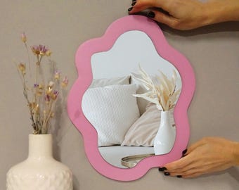May include: A pink cloud-shaped mirror with a wavy edge. The mirror reflects a white pillow and a vase with dried flowers.