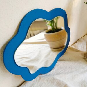 May include: A blue, organically shaped mirror is mounted on a white wall. The mirror's frame is a bright blue, and the reflection shows a room with plants. The mirror's unusual shape adds a contemporary feel to the decor.