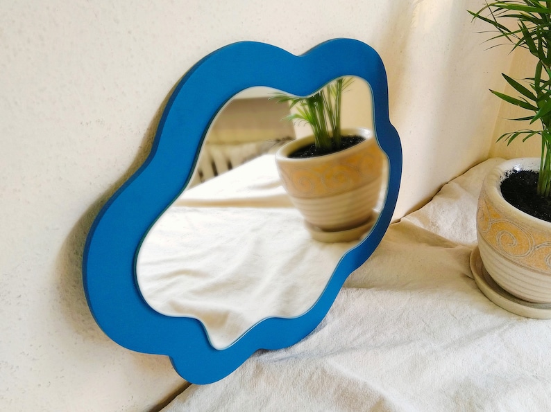 May include: A blue, organically shaped mirror is mounted on a white wall. The mirror's frame is a bright blue, and the reflection shows a room with plants. The mirror's unusual shape adds a contemporary feel to the decor.