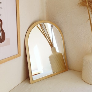 May include: A wooden-framed, arched mirror leans against a neutral-colored wall. The mirror reflects a white vase with a textured surface, filled with dried reeds. A framed artwork is visible on the left. The overall aesthetic is minimalist and natural.