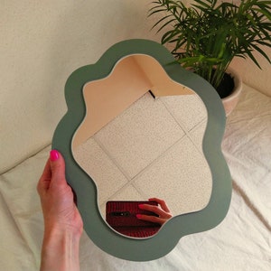 May include: A green, wavy-shaped mirror with a white ceiling reflected in the glass. The mirror is being held by a hand with pink nail polish.