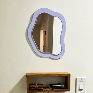 May include: A modern, wavy-edged mirror with a light purple frame hangs on a white wall. Below, a wooden shelf with a wallet and keys is mounted. A light switch is visible to the right. The mirror's unique shape adds a contemporary touch to the space.