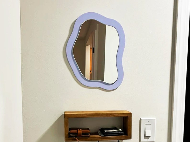 May include: A modern, wavy-edged mirror with a light purple frame hangs on a white wall. Below, a wooden shelf with a wallet and keys is mounted. A light switch is visible to the right. The mirror's unique shape adds a contemporary touch to the space.