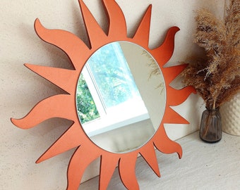 May include: A terracotta-colored sunburst mirror with a round, reflective surface. The mirror is made of wood and has a rustic, bohemian style.