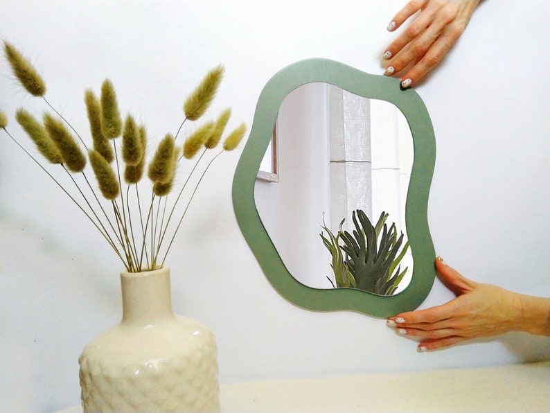 May include: A sage green, organically shaped wall mirror. The mirror is held against a white wall, reflecting a glimpse of greenery. A vase with dried, fluffy wheat-like stems sits to the left.