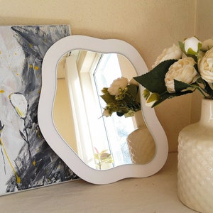 May include: A white, wavy-edged mirror reflects a window and curtains. The mirror is set on a white surface, next to a vase of white flowers and a painting with black, white, and yellow brushstrokes. The mirror's frame has a smooth finish.