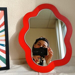 May include: A red, flower-shaped mirror with a wavy edge. The mirror is hanging on a white wall and reflects a person taking a selfie.