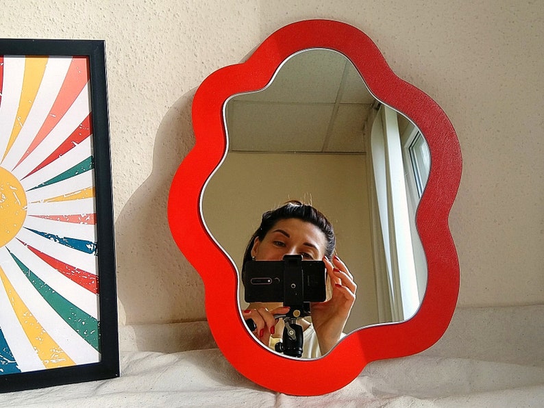 May include: A red, flower-shaped mirror with a wavy edge. The mirror is hanging on a white wall and reflects a person taking a selfie.
