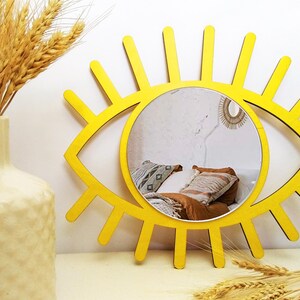 K&ouml;nnte beinhalten: Ein leuchtend gelber, augenf&ouml;rmiger Wandspiegel mit einer kreisf&ouml;rmigen Glasmitte. Der Rahmen besteht aus strahlenf&ouml;rmig angeordneten Holzlatten, die an Wimpern erinnern. Der Spiegel reflektiert ein Schlafzimmer mit Kissen und einem kleinen Wandbehang.