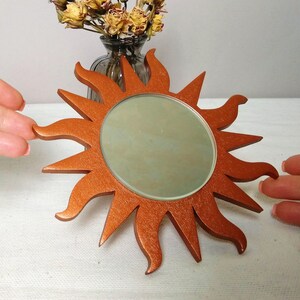 May include: A copper-colored sunburst mirror with a round mirror in the center. The mirror is surrounded by a decorative sunburst design.