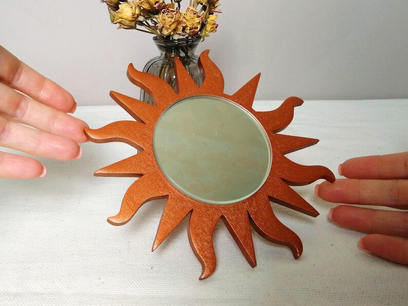 May include: A copper-colored sunburst mirror with a round mirror in the center. The mirror is surrounded by a decorative sunburst design.