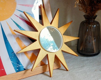 May include: A gold-colored starburst mirror with a round mirror in the center. The starburst has pointed rays extending outwards. The mirror is placed in front of a framed print with a sun design and colorful stripes.