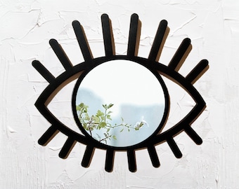 Könnte beinhalten: Schwarzer, augenförmiger Spiegel mit langen Wimpern, der einen Blick auf grüne Pflanzen und weiße Blumen reflektiert.