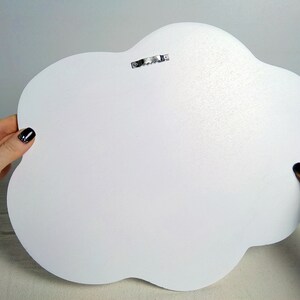 May include: A white cloud-shaped wooden wall decoration with a metal sawtooth hanger on the back. The cloud has a smooth, matte finish. The image shows the back of the cloud, held by hands with dark nail polish.